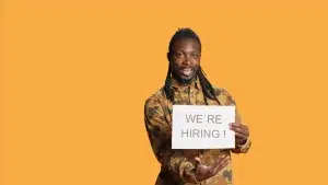 Man in camouflage overhemd houdt een bord vast met de tekst "We’re hiring", symbool voor professioneel inhuren van externe arbeid.